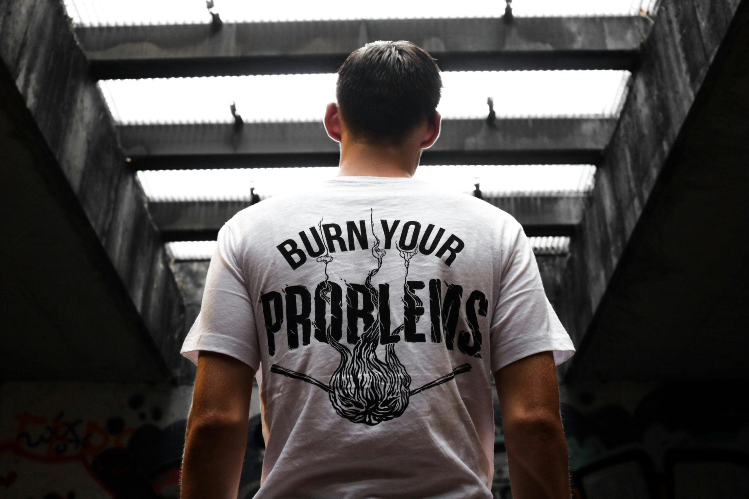 Man standing indoors, wearing a t-shirt with 'Burn Your Problems' text, viewed from behind.